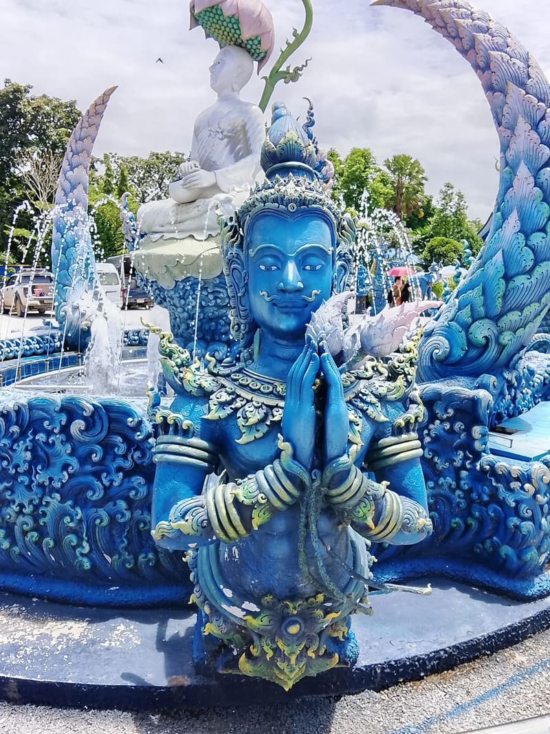 Blue statue of a deva in front of the temple, in a 'wai' posture to greet and protect.