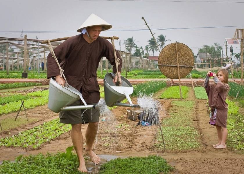A Western man waters garden beds using traditional metal cans in Hoi An, Vietnam.